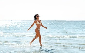 people, summer and swimwear concept - young woman in bikini swimsuit running in shallow water on beach. young woman in bikini swimsuit running on beach