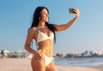 people, summer and leisure concept - happy smiling young woman in bikini swimsuit taking selfie with smartphone on beach. smiling woman in bikini taking selfie on beach