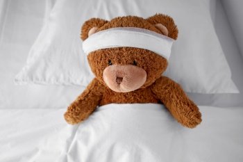 medicine, healthcare and childhood concept - ill teddy bear toy with bandaged head lying in bed. ill teddy bear toy with bandaged head lying in bed