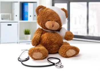 medicine, healthcare and childhood concept - teddy bear toy with bandaged head and paw and stethoscope over medical office at hospital background. bandaged teddy bear toy with stethoscope