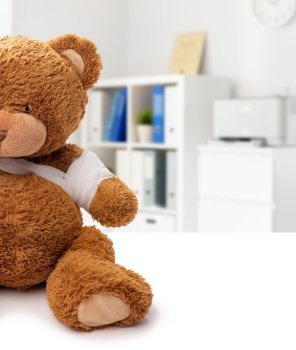 medicine, healthcare and childhood concept - teddy bear toy with bandaged paw over medical office at hospital background. teddy bear toy with bandaged paw