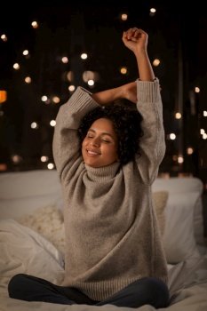 winter, comfort and people concept - happy smiling woman in woolen high neck sweater stretching in bed at home at night. woman in sweater stretching in bed at night