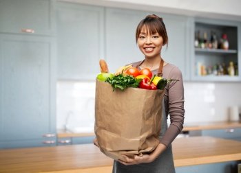 cooking, culinary and people concept - happy smiling woman in apron holding food in paper bag over home kitchen background. smiling woman with food in paper bag at kitchen