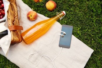 leisure, technology and summer concept - close up of picnic basket, bottle of fruit juice and smartphone with earphones on grass. phone, earphones, picnic basket and juice bottle