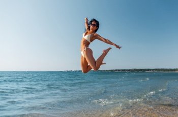 people, summer and swimwear concept - happy smiling young woman in bikini swimsuit jumping on beach. smiling young woman in bikini swimsuit on beach