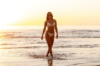 people, summer and leisure concept - happy smiling young woman in bikini swimsuit on beach over sunset. happy smiling woman in bikini on beach over sunset