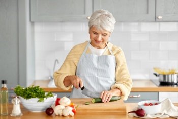 healthy eating, food cooking and culinary concept - happy smiling senior woman with knife chopping cucumber on kitchen at home. happy woman chopping cucumber on kitchen