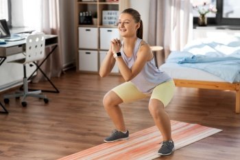 sport, fitness and healthy lifestyle concept - smiling teenage girl with smart watch exercising on yoga mat and doing squats at home. happy teenage girl exercising on yoga mat at home