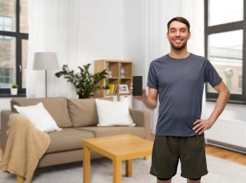 fitness, sport and healthy lifestyle concept - smiling man in sports clothes showing smartphone over home room background. smiling man in sports clothes showing smartphone