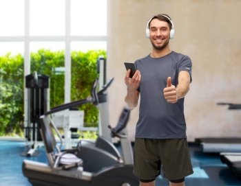 fitness, sport and healthy lifestyle concept - smiling man in sports clothes with smartphone and headphones listening to music and showing thumbs up over gym background. man with phone and headphones in gym