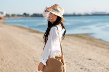 people, summer holidays and leisure concept - happy smiling woman in white shirt and straw hat with jute bag walking along beach. happy woman with bag walking along summer beach