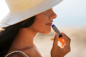 people, summer and swimwear concept - close up of happy smiling young woman in bikini swimsuit and straw hat applying lip balm on beach. smiling woman in bikini with lip balm on beach