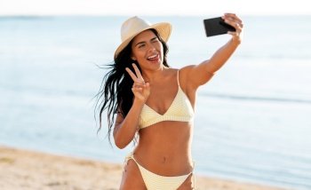 people, summer and swimwear concept - happy smiling young woman in bikini swimsuit and straw hat taking selfie with smartphone on beach. smiling woman in bikini taking selfie on beach