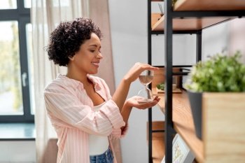 home improvement and people concept - happy smiling woman holding hourglass at shelf. smiling woman with hourglass at shelf at home