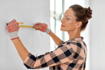 repair, construction and building concept - woman in goggles and protective gloves with ruler and pencil measuring wall at home. woman with ruler and pencil measuring wall at home