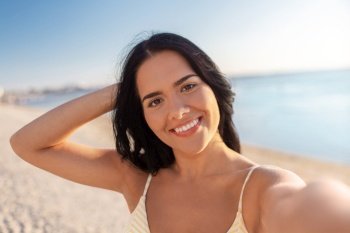 people, summer and swimwear concept - happy smiling young woman in bikini swimsuit taking selfie on beach. smiling woman in bikini taking selfie on beach