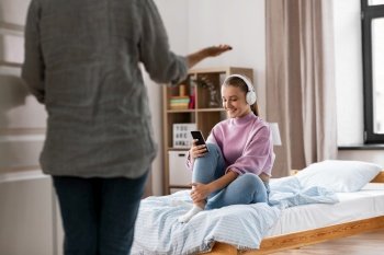 misbehavior, conflict and family concept - angry mother entering room and smiling daughter in headphones with smartphone sitting on bed at home. angry mother and daughter with smartphone at home