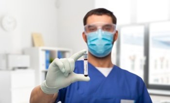 healthcare, coronavirus and medicine concept - doctor in blue uniform, face protective medical mask, goggles and gloves with blood in test tube over medical office at hospital background. male doctor in mask with blood in test tube