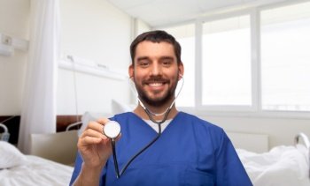 healthcare, profession and medicine concept - happy smiling doctor or male nurse in blue uniform with stethoscope over hospital ward background. smiling doctor or male nurse with stethoscope