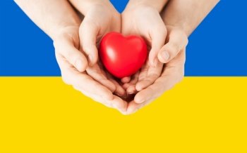 national, patriotic and charity concept - close up of woman and man hands holding red heart shape over colors of flag of ukraine on background. hands holding red heart shape over flag of ukraine