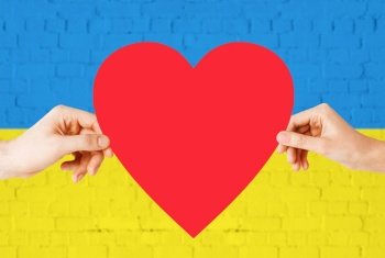 national, patriotic and charity concept - close up of woman and man hands holding red heart shape over brick wall painted in colors of flag of ukraine. hands holding red heart shape over flag of ukraine