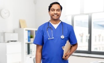 healthcare, profession and medicine concept - happy smiling doctor or male nurse in blue uniform with clipboard and stethoscope over medical office at hospital background. male doctor with clipboard and stethoscope