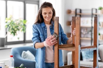 furniture renovation, diy and home improvement concept - happy smiling woman sanding old wooden table or chair with sponge. woman sanding old round wooden table with sponge