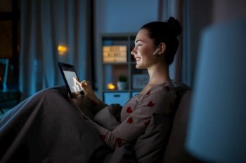 technology, bedtime and people concept - happy smiling teenage girl with tablet pc computer and earphones sitting in bed at home at night. teenage girl with tablet pc and earphones in bed