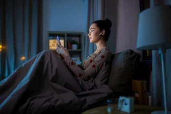 technology, bedtime and people concept - teenage girl with smartphone and earphones sitting in bed at home at night. teenage girl with phone and earphones in bed
