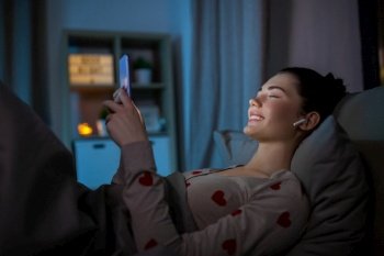 technology, bedtime and people concept - happy teenage girl with smartphone and earphones enjoying music lying in bed at home at night. happy teenage girl with phone and earphones in bed