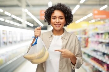 sustainability, shopping and food concept - portrait of happy smiling woman holding reusable string bag with bananas over supermarket or grocery store background. happy woman with bananas in reusable string bag