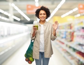 sustainability, shopping and people concept - happy smiling young woman with food in reusable string bag drinking green takeaway smoothie or shake over supermarket or grocery store background. woman with reusable shopping bag drinking smoothie