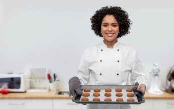 cooking, culinary and bakery concept - happy smiling female chef or baker in white jacket holding baking tray with oatmeal cookies over restaurant kitchen background. happy female chef with cookies on oven tray