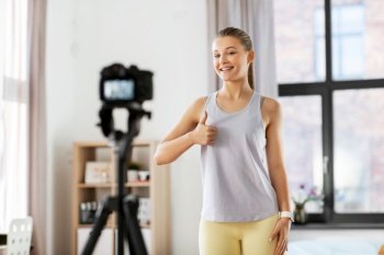 fitness, sport and video blogging concept - happy smiling teenage girl or blogger with camera on tripod recording online gym class at home and showing thumbs up. teenage girl or blogger recording gym class video