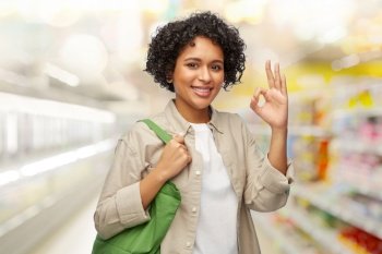 eco living, zero waste and sustainability concept - portrait of happy smiling woman with green reusable canvas bag for food shopping showing ok gesture over supermarket background. woman with reusable canvas bag for food shopping