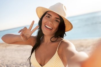 people, summer and swimwear concept - happy smiling young woman in bikini swimsuit and straw hat taking selfie on beach and showing peace gesture. smiling woman in bikini taking selfie on beach