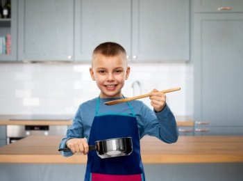 culinary and profession concept - happy smiling little boy in apron with saucepan and wooden spoon cooking food over kitchen background. little boy in apron with saucepan cooking food