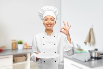 cooking, culinary and people concept - happy smiling female chef holding empty plate showing ok gesture over kitchen background. female chef holding empty plate and showing ok