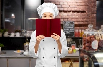 cooking, culinary and people concept - female chef hiding behind cook book over restaurant kitchen background. female chef hiding behind cook book