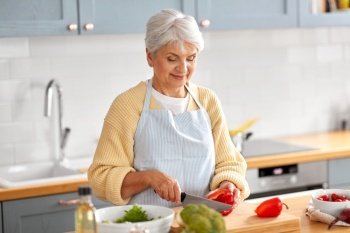 healthy eating, food cooking and culinary concept - happy smiling senior woman with knife chopping red pepper on kitchen at home. happy woman chopping red pepper on kitchen