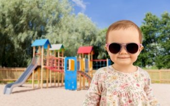 childhood, summer and people concept - happy little baby girl in sunglasses over children’s playground background. happy baby girl in sunglasses over playground