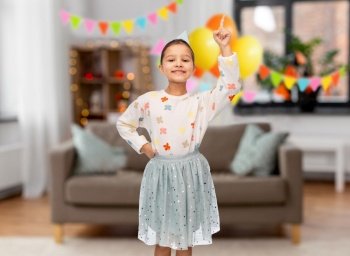 birthday, childhood and people concept - portrait of smiling little girl in party hat pointing finger up over decorated living room background. happy girl in birthday cap pointing finger up