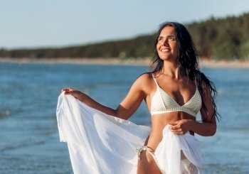 people, summer and swimwear concept - happy smiling young woman in bikini swimsuit with pareo posing on beach. woman in bikini swimsuit with pareo on beach