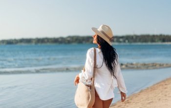 people, summer holidays and leisure concept - happy young woman in white shirt and straw hat with bag walking along beach. happy woman with bag walking along summer beach