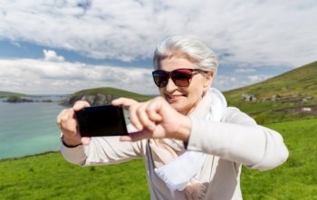 travel, tourism and vacation concept - happy smiling senior woman taking selfie by smartphone over atlantic ocean coast in ireland background. senior woman taking selfie by smartphone on beach