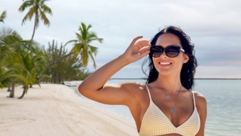 travel, tourism and summer vacation concept - happy smiling young woman in sunglasses and bikini swimsuit over tropical beach background in french polynesia. smiling young woman in sunglasses on summer beach