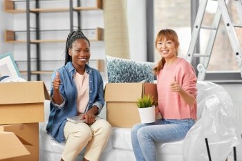 moving, people and real estate concept - women unpacking boxes and showing thumbs up gesture at new home. women unpacking boxes and moving to new home