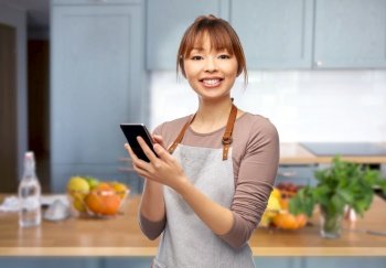 cooking, technology and people concept - happy smiling female chef or waitress in apron showing smartphone with empty screen over home kitchen background. happy woman in apron showing smartphone in kitchen