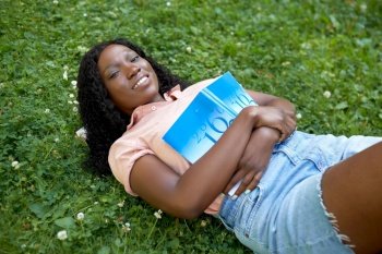 education, school and people concept - happy smiling african american student girl with math textbook lying on grass. african student girl with textbook lying on grass