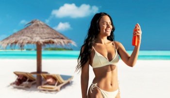 people, summer and swimwear concept - happy smiling young woman in bikini swimsuit using sunscreen spray over palapa and sun beds on tropical beach background in french polynesia. smiling woman in bikini with sunscreen on beach